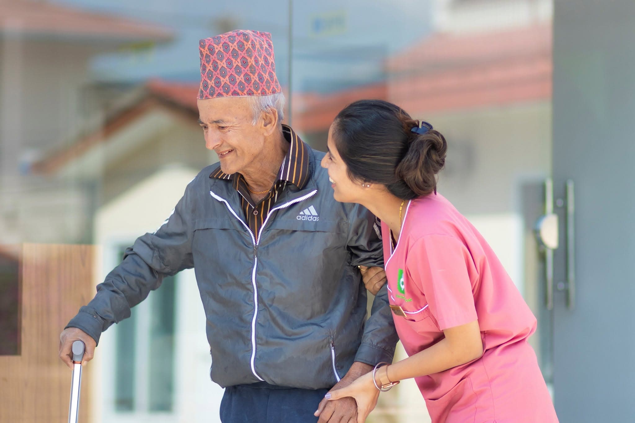 A Naman nurse walking arm-in-arm with an elderly resident in a Nepali dhaka topi — everyday elderly living